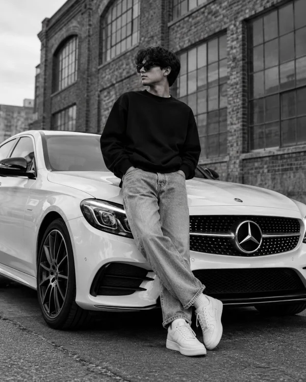 Black and white AI fashion portrait of a stylish young man wearing sunglasses and streetwear while leaning against a white Mercedes-Benz luxury car.